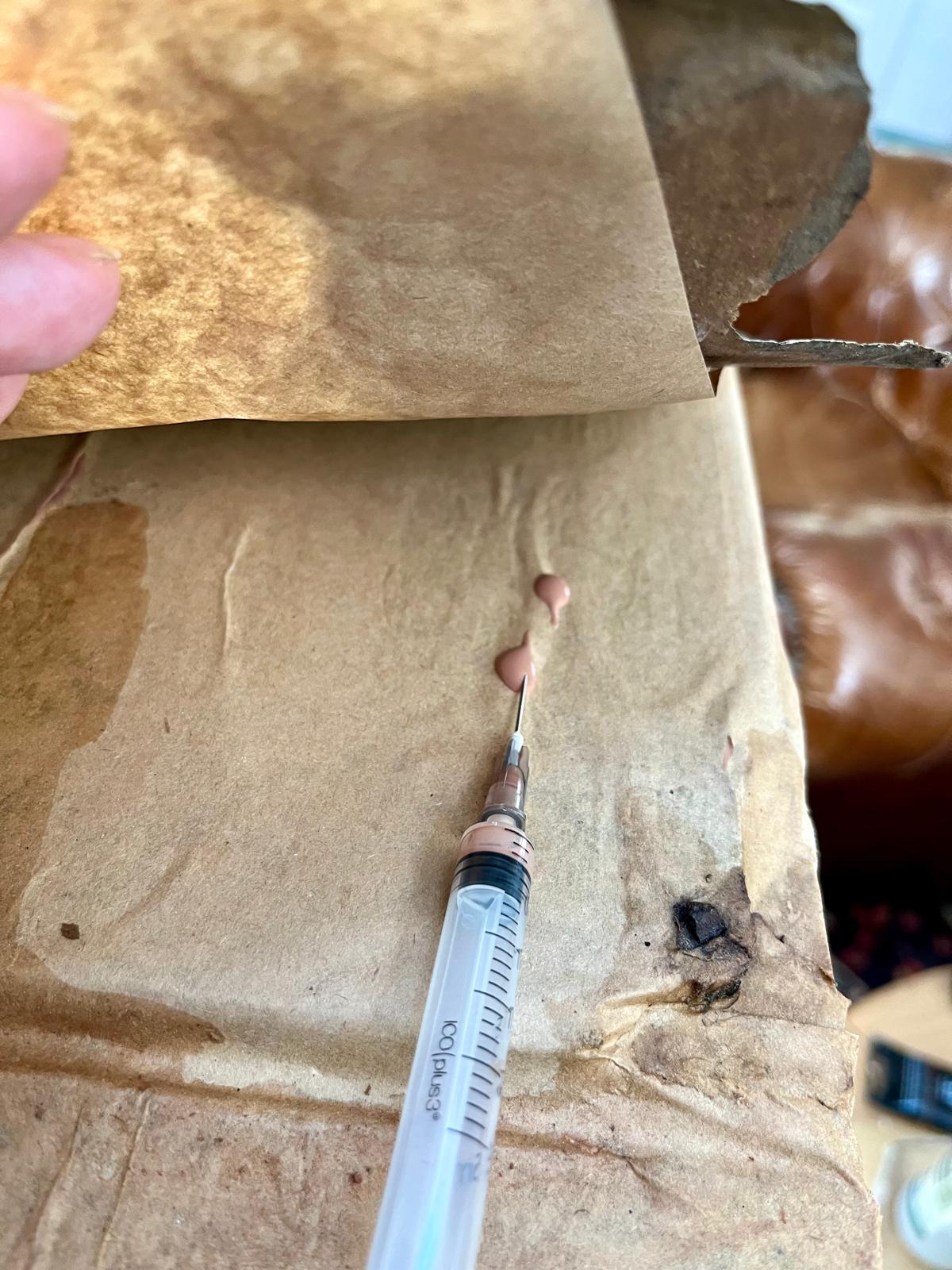 A hand lifting the edge of the antique paper, the syringe needle with a drop of the compound ready to inject